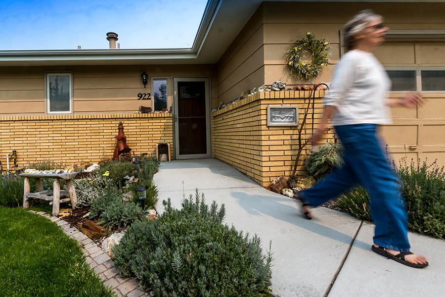 A woman with blue jeans, a grey top and sunglasses is photographed mid-stride walking even farther away from the entrance to the home.